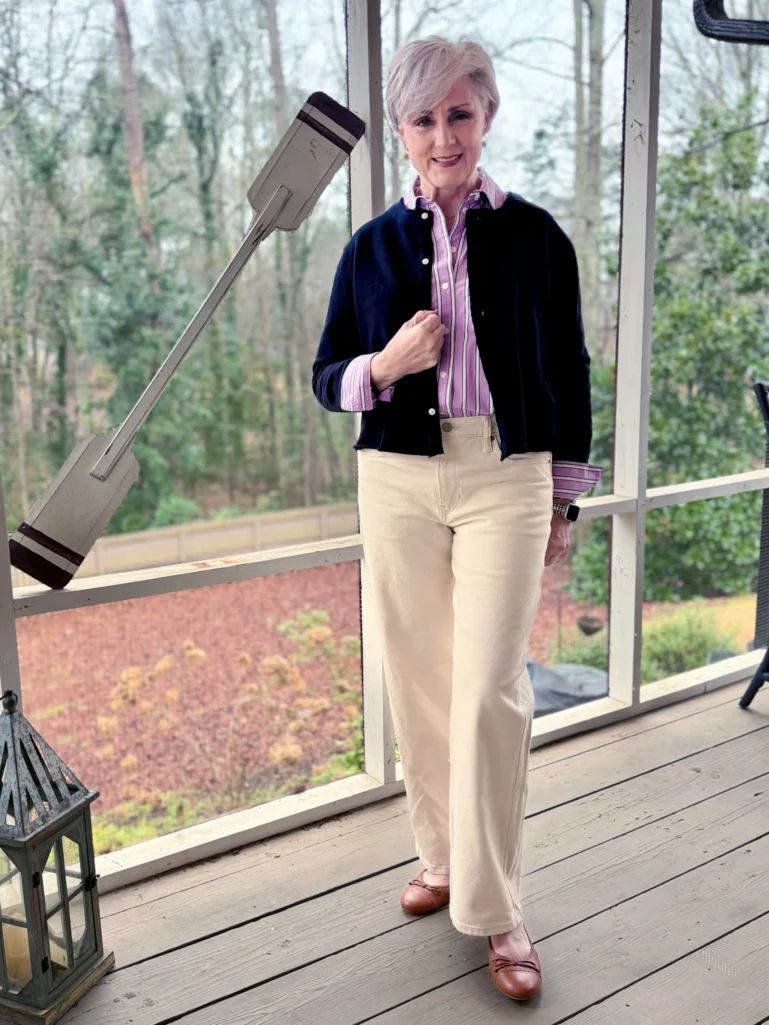 Woman over 50 wearing a pink and purple vertical striped button-up shirt layered under a navy cardigan with cream wide-leg pants and brown ballet flats, demonstrating how to style vertical stripes for an elongating effect.