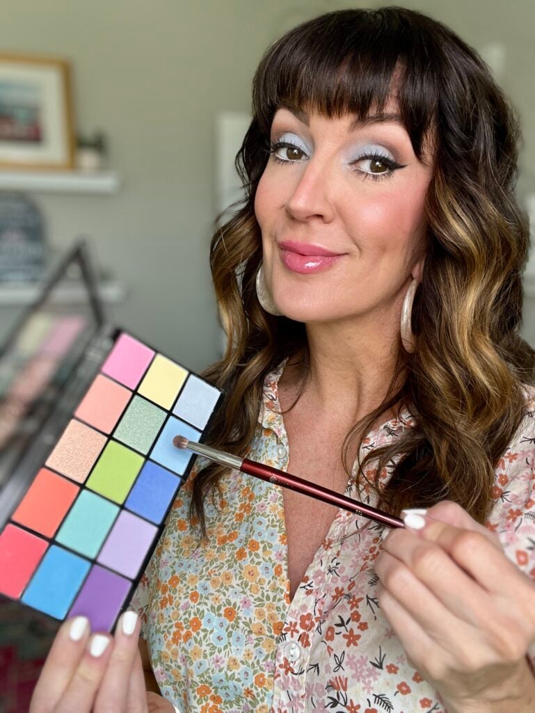 Woman holding colorful rainbow eyeshadow palette with pastel and bright shades and burgundy eyeshadow brush, wearing blue and lavender eyeshadow look on lids, pastel floral print blouse, large gold teardrop hoop earrings, glossy pink lips, white manicure