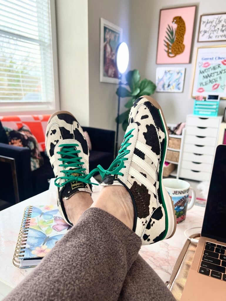 Adidas SL 72 OG sneakers in cow print pony hair with green laces and green sole trim, ivory and chocolate brown pattern, feet propped on desk next to butterfly spiral notebook, personalized coffee mug, laptop edge visible, colorful gallery wall in home office background