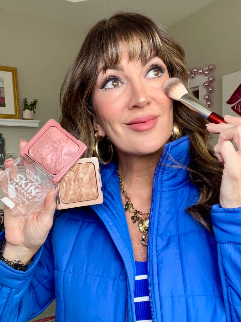 Woman in royal blue puffer jacket holding CoverGirl TruBlend Skin Enhancer blush in pink case and bronzer in nude case while pressing a makeup brush to cheek, playful expression, gold hoop earrings and layered charm necklace