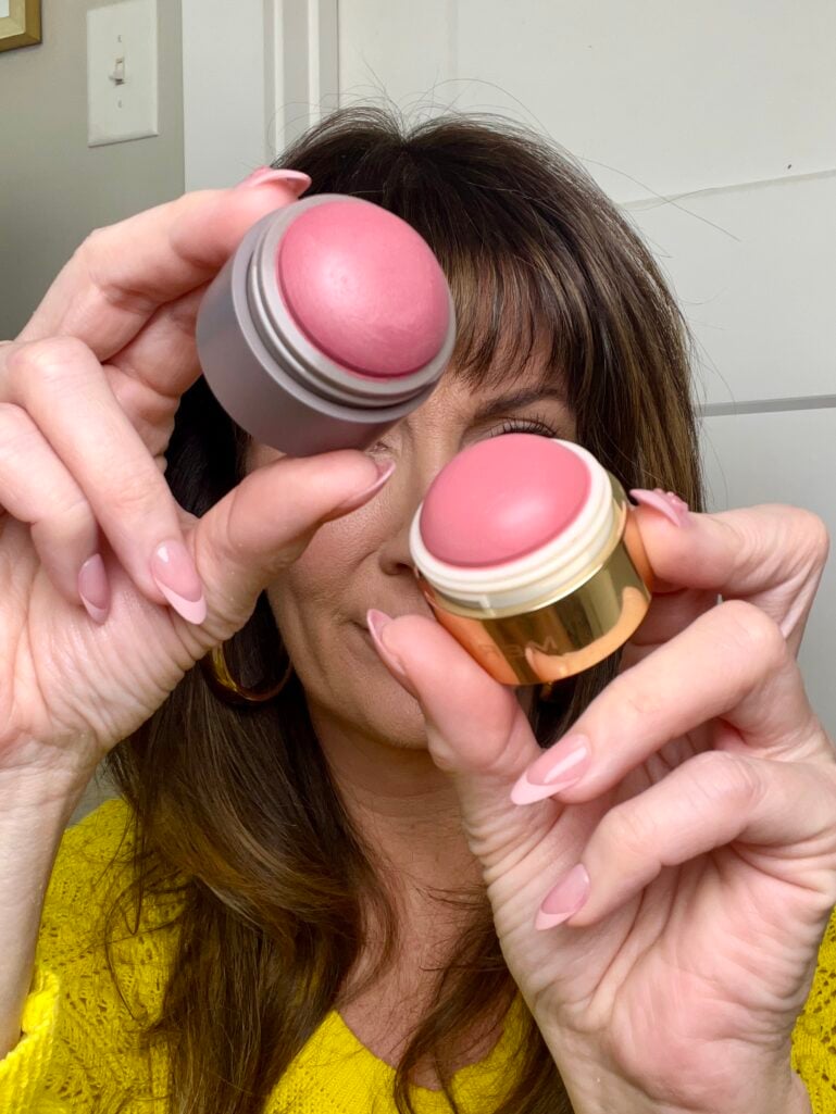 Woman holding two Merit Flush Balm cream blushes up to eyes—one in gray packaging and one in gold packaging—showing the rounded dome blush formula in rosy pink shade Cheeky, wearing yellow sweater, gold hoop earrings partially visible, pink French tip nails