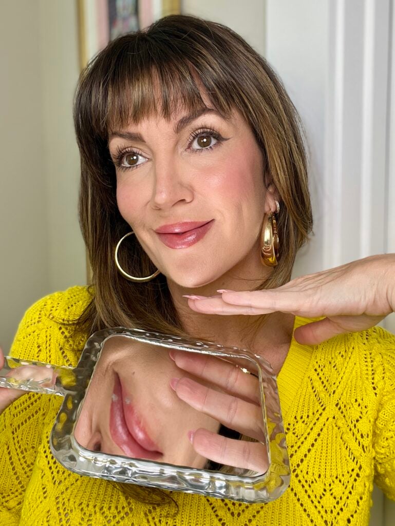 Woman with completed foundation look holding wavy glass mirror showing reflection of her chin and lips, wearing yellow pointelle knit sweater, gold puffy hoop earrings, luminous radiant skin, rosy flushed cheeks, mauve-pink lips, long lashes, pink French tip nails