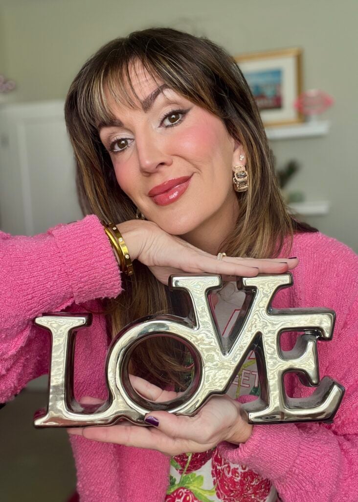 Woman with highlighted brown hair holding silver metallic LOVE sign decoration, wearing pink textured cardigan over strawberry print tee, romantic makeup with rosy cheeks, glossy rose lips, smokey eyes, gold chunky hoop earrings, Cartier-style gold bangle, and dark purple nail polish