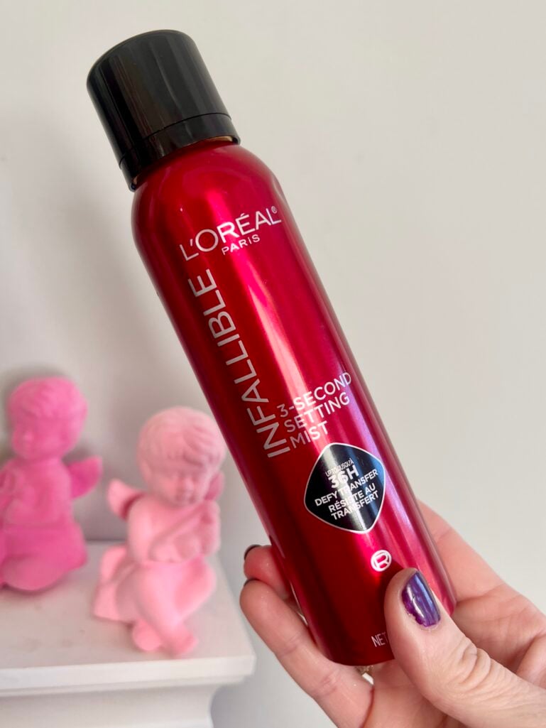 Hand holding red L'Oreal Infallible 3-Second Setting Mist spray bottle with black cap, pink flocked cupid figurines in background on white shelf, dark purple nail polish visible