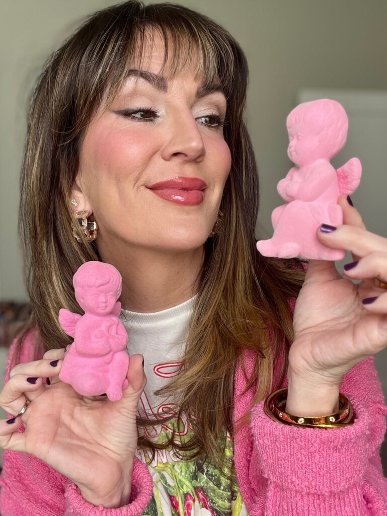 Woman holding two pink flocked cupid figurines, wearing pink textured cardigan over strawberry print tee, romantic Valentine's makeup with flushed cheeks and glossy rose lips, gold chunky hoop earrings with crystal detail, gold bangle bracelet, dark purple nails
