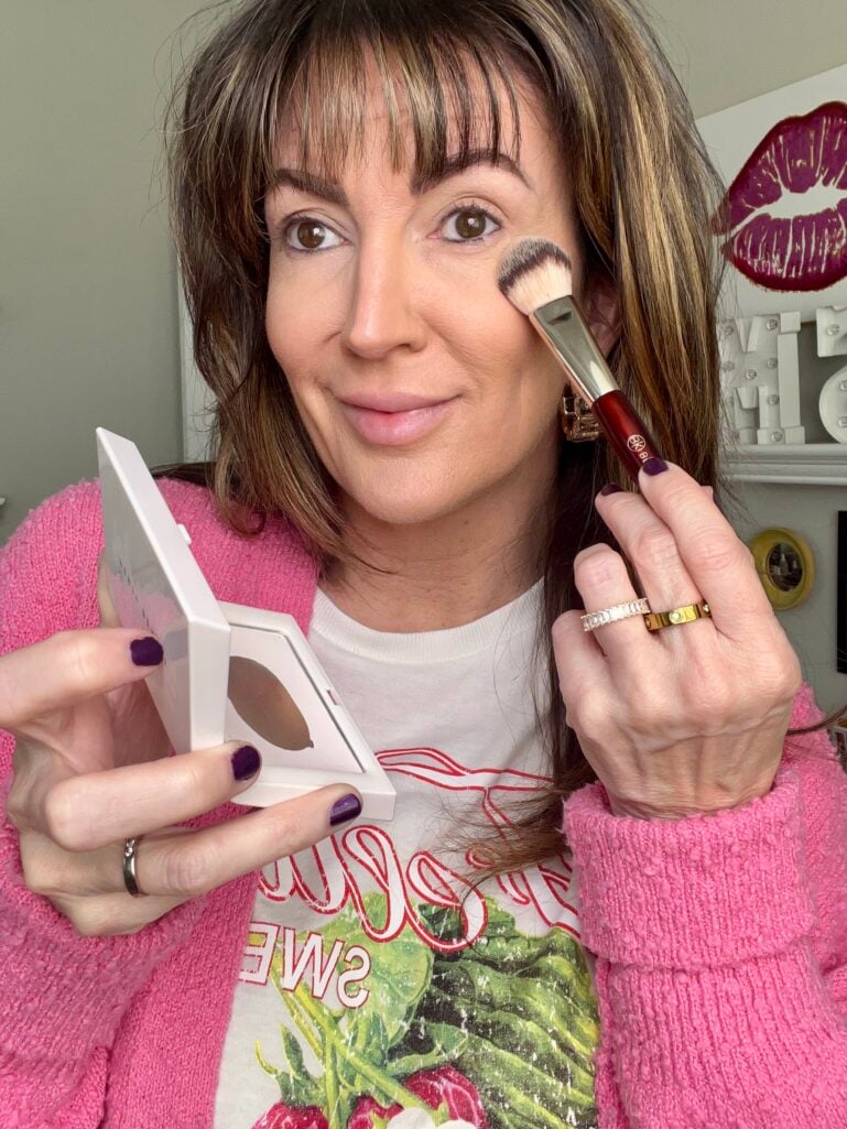 Woman applying Fenty Beauty Invisimatte setting powder to cheek with BK Beauty brush while holding open white compact with mirror, wearing pink cardigan over strawberry print tee, natural skin prep visible, gold rings including Cartier-style band, dark purple nails