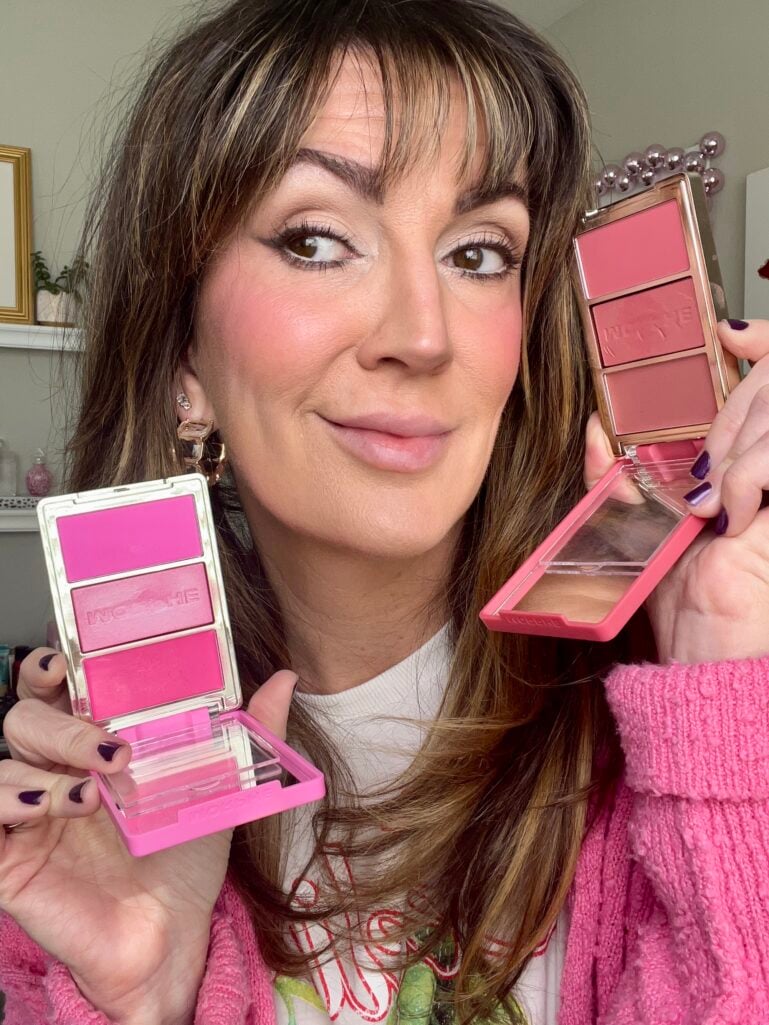 Woman holding two open Morphe Cheek Thrills blush trio palettes in shades Blind Date (bright pink) and Apres Ski (dusty rose), wearing pink cardigan, gold hoop earrings, soft smokey eye makeup with rosy flushed cheeks, dark purple nail polish