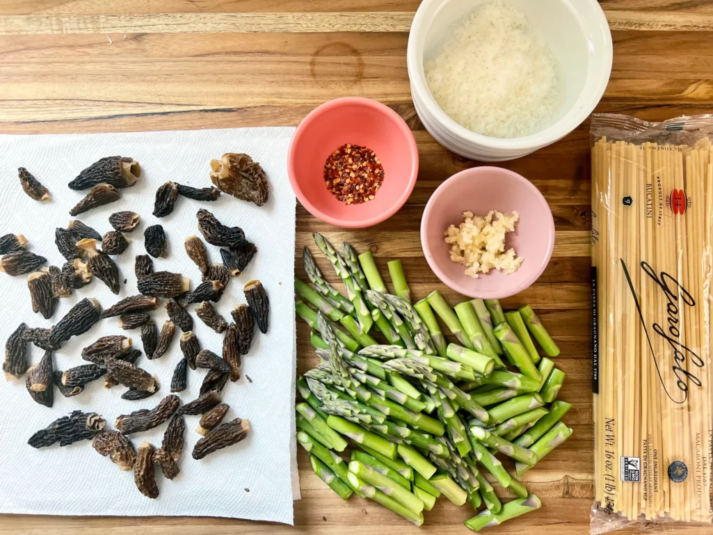 Prepped ingredients on a wooden cutting board: reconstituted morels patted dry on paper towels, asparagus cut into pieces, minced garlic in a pink bowl, grated Parmesan in a white bowl, crushed red pepper, and bucatini