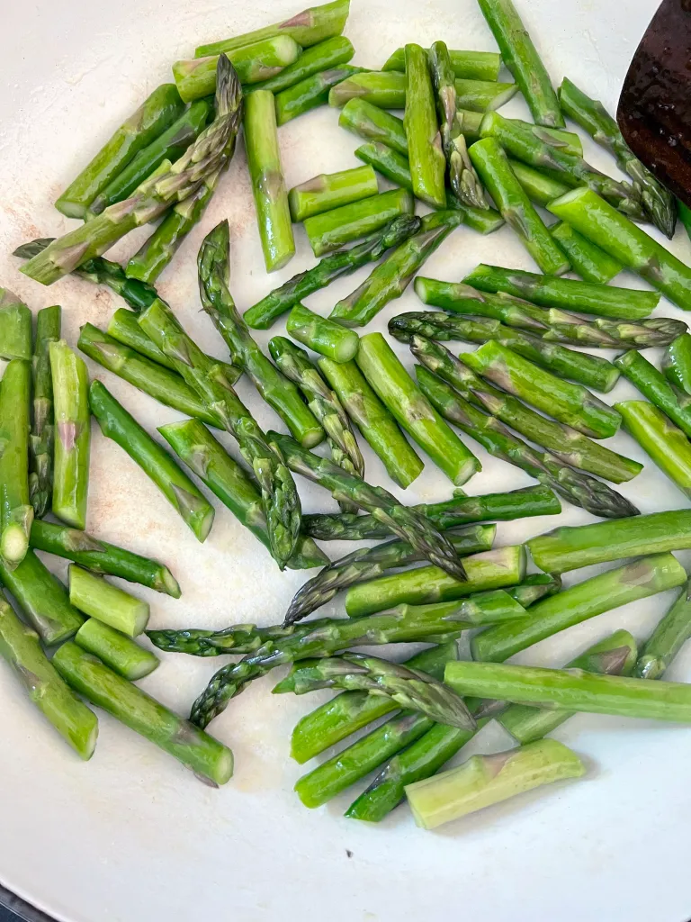 Bright green asparagus pieces sautéing in olive oil in a white skillet with a wooden spoon