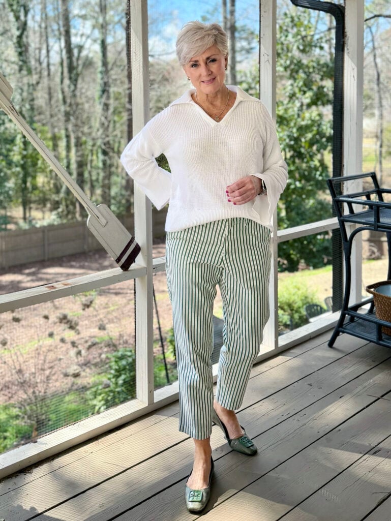 Beth wearing a white collared v-neck sweater, green and white striped mid-rise classic straight leg chino pants, and green snake-embossed flats on the screened porch