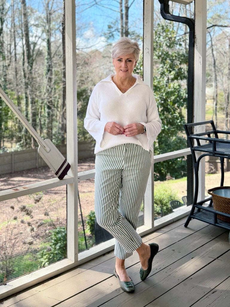 Full-length view of Beth in her white collared sweater and green striped chinos, hands together, spring greenery visible through the porch screens