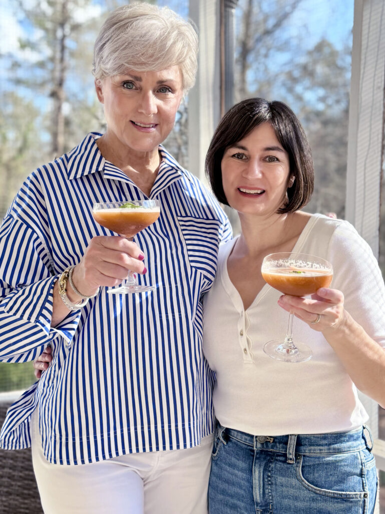 Beth and Kelly holding Strawberry Basil Smash cocktails in coupe glasses, the drinks a beautiful deep strawberry-red with basil leaf garnish, Beth in her striped popover and Kelly in her white ribbed henley