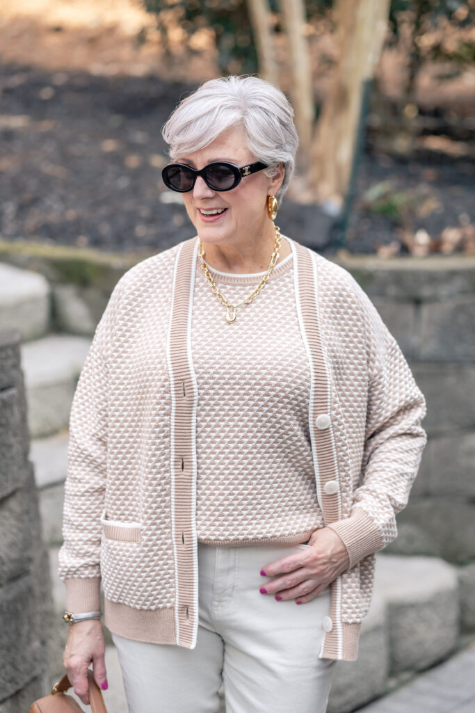 Close-up of woman over 50 with silver hair wearing a chunky gold chain link necklace with a gold pendant and oversized gold dome earrings, styled with a cream and taupe textured knit cardigan and matching sweater — chunky gold statement jewelry for women over 50, spring 2026