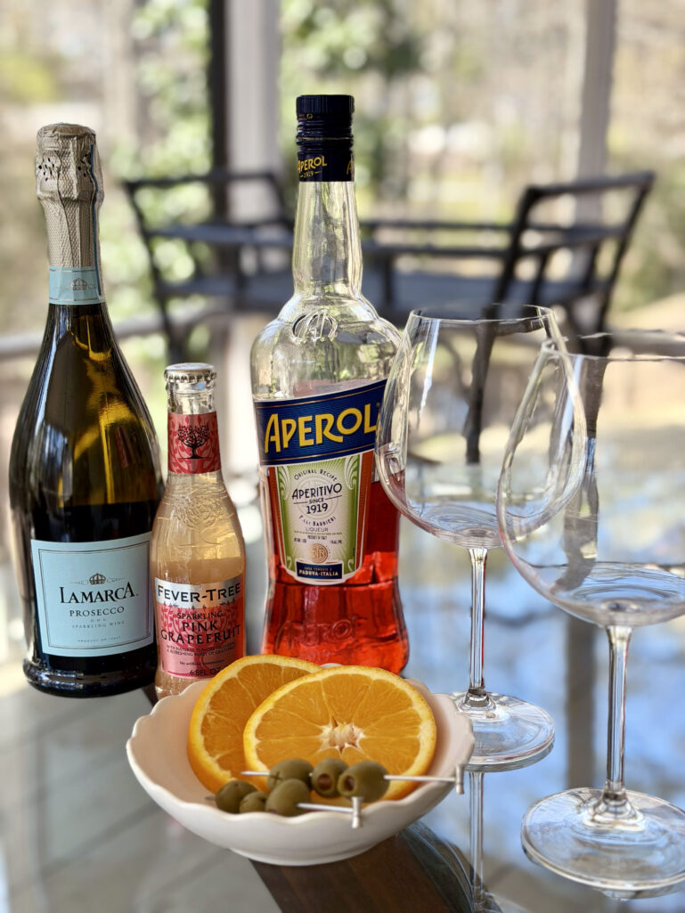 Aperol Spritz ingredients on a glass table: a bottle of La Marca Prosecco, a small bottle of Fever-Tree Sparkling Pink Grapefruit, a bottle of Aperol, two large empty wine glasses, and a small bowl of sliced oranges and green olives on cocktail picks.