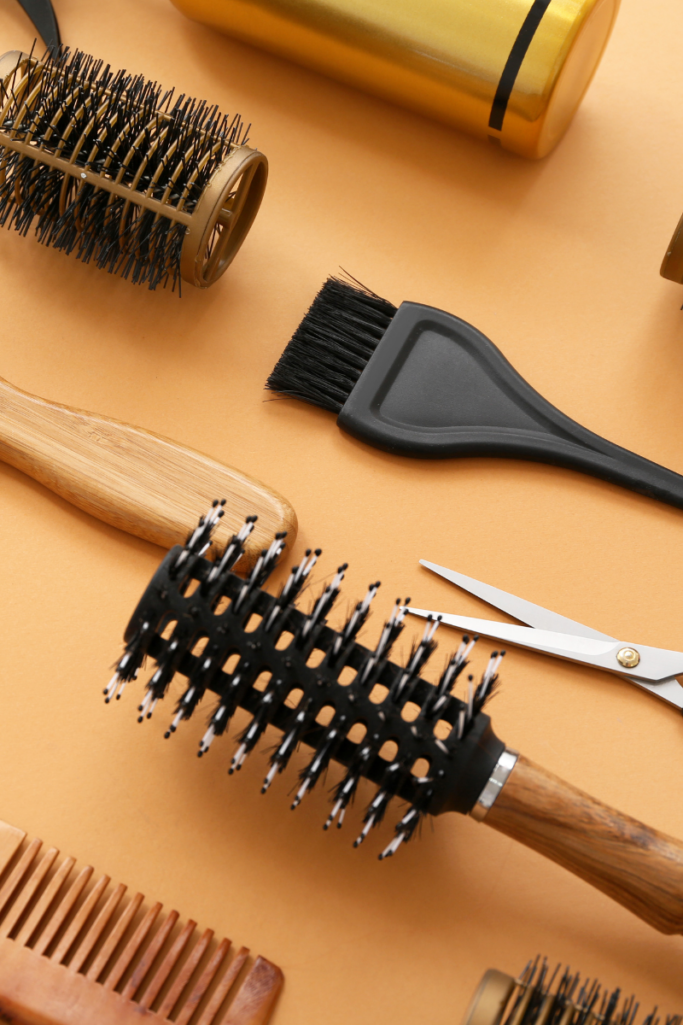 Flat lay of hair styling tools including round brushes, a tint brush, combs, scissors, and a gold styling tool, arranged on a warm neutral background for a clean, salon-inspired look.