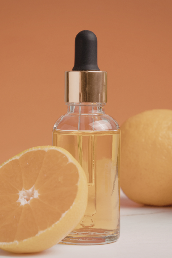 Glass dropper bottle filled with golden vitamin C serum beside a sliced grapefruit and whole citrus fruit against a warm peach background, representing brightening skincare ingredients.
