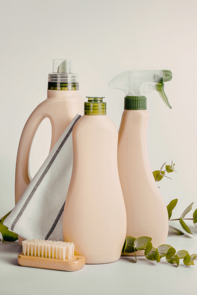 Minimalist cleaning setup featuring neutral-toned reusable spray bottles, a wooden scrub brush, folded cleaning cloth, and eucalyptus stems arranged on a white surface.