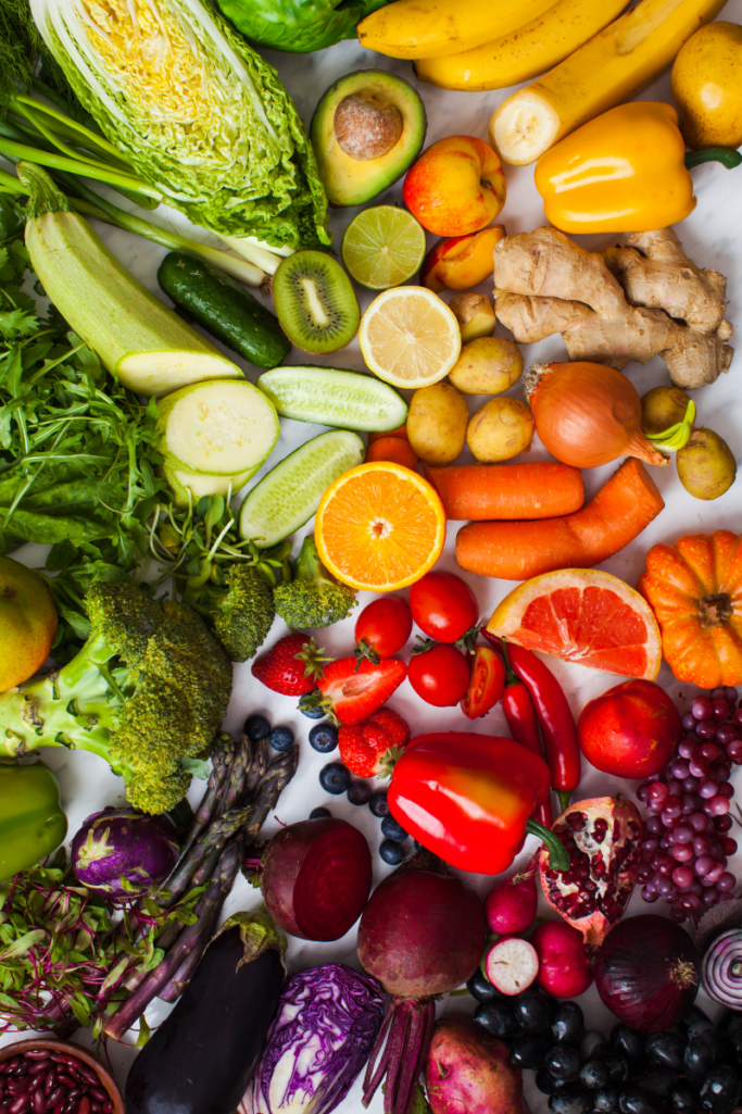 Colorful assortment of fresh fruits and vegetables arranged on a white surface, including broccoli, carrots, citrus, bell peppers, cabbage, grapes, berries, leafy greens, and root vegetables.