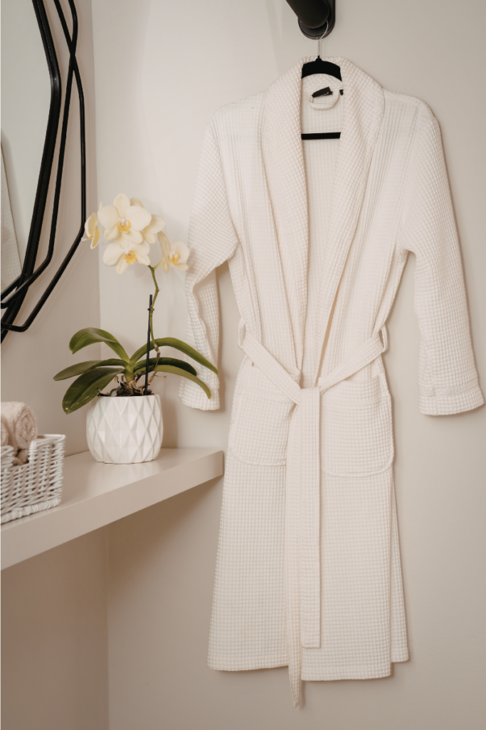White waffle robe hanging on a wall hook in a serene bathroom setting, styled beside a potted orchid, folded towels, and a minimalist shelf for a clean, spa-like feel.