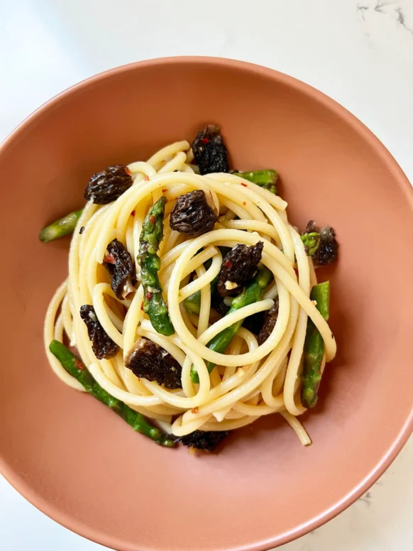 Overhead view of plated bucatini with asparagus and morel mushrooms in a terracotta bowl on a marble countertop