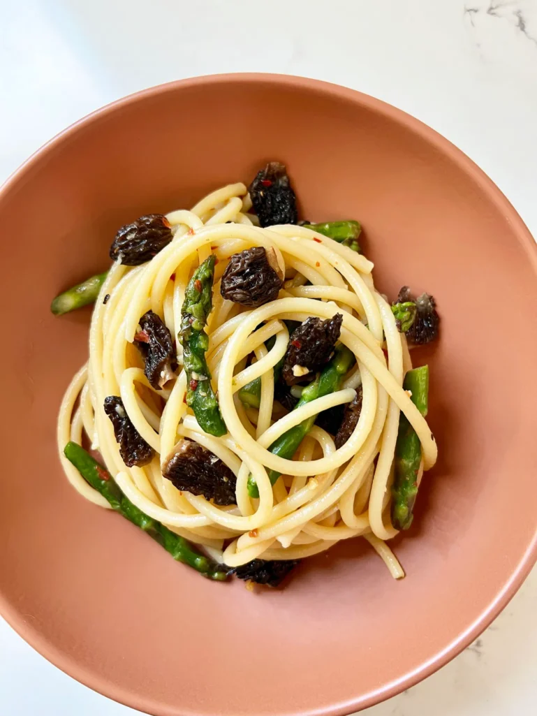 Overhead view of plated bucatini with asparagus and morel mushrooms in a terracotta bowl on a marble countertop