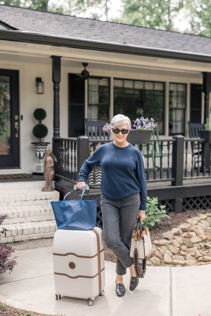 Woman over 50 with silver hair wearing a navy crewneck sweater with dark wash straight-leg jeans and black horsebit loafers, leaning on a cream and cognac hardshell spinner suitcase while carrying a blue woven tote and a black and cream shoulder bag in front of a white and black farmhouse porch — Amazon Big Spring Sale travel must-haves for women over 50