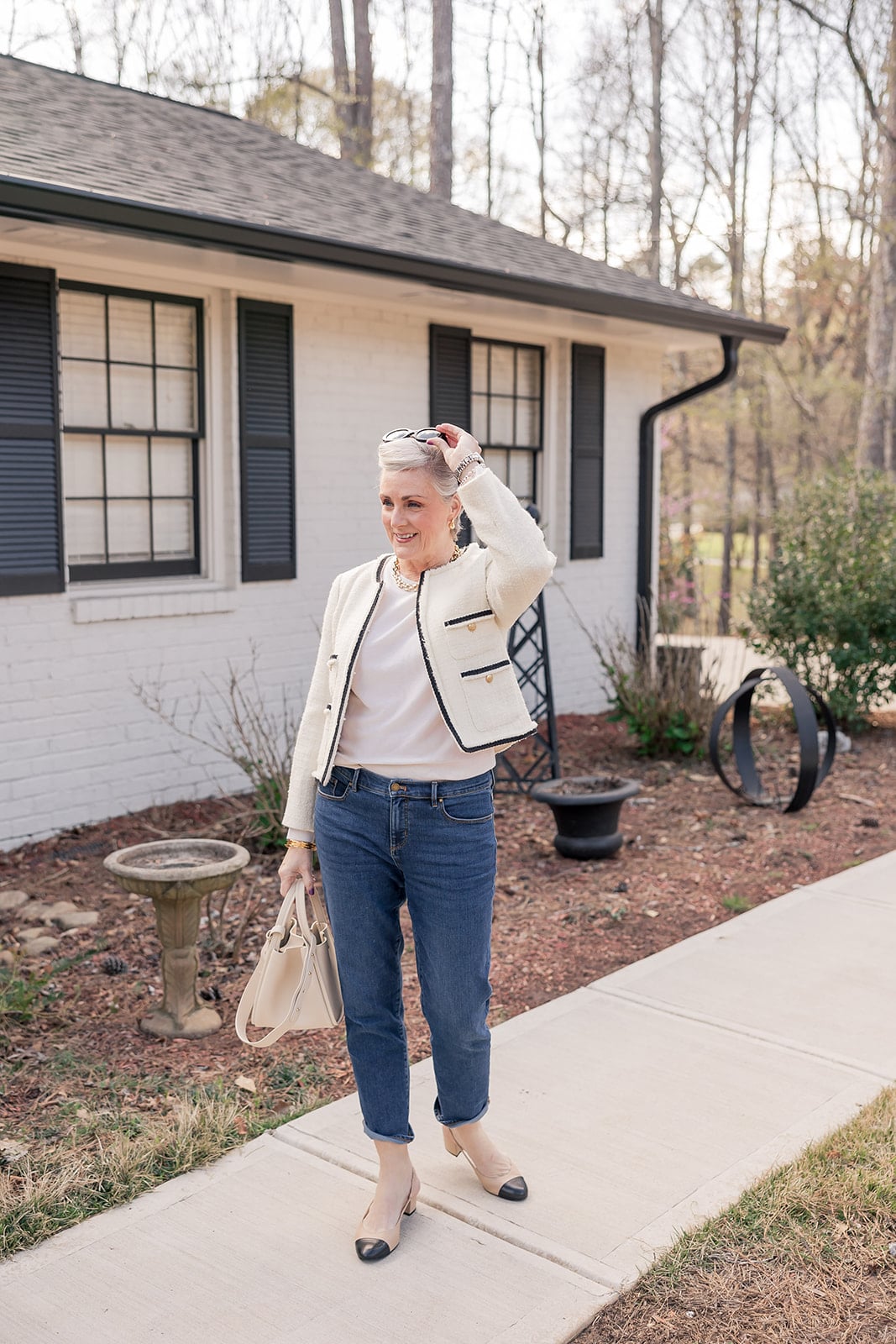 Beth wearing a cream tweed jacket with black trim and gold buttons, white tee, cuffed straight-leg jeans, and beige and black cap-toe block heel slingbacks, carrying a cream structured top-handle bag and wearing a gold chain necklace and black sunglasses