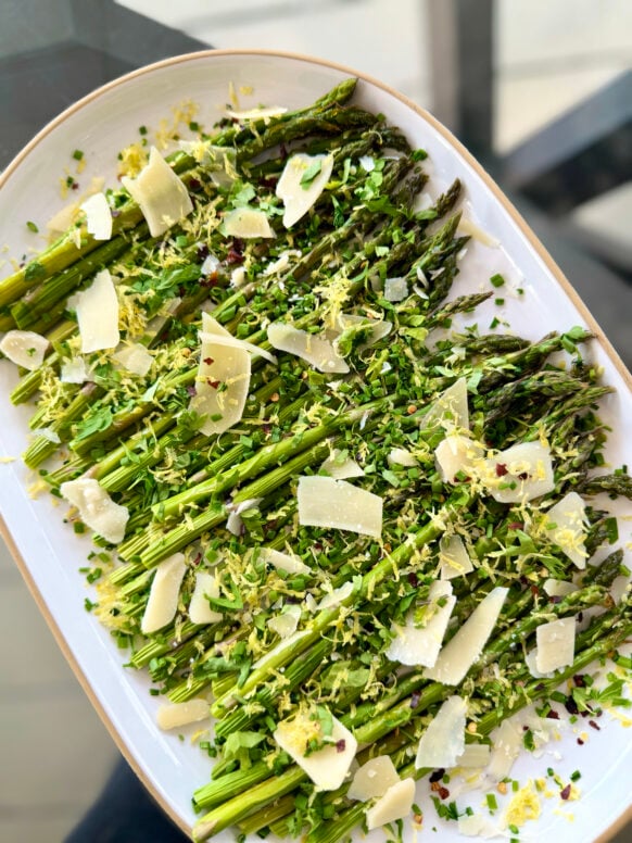Overhead view of Roasted Asparagus with Shaved Parmesan on a white oval platter, topped with lemon zest, fresh chives, red pepper flakes, and wide Parmesan shavings