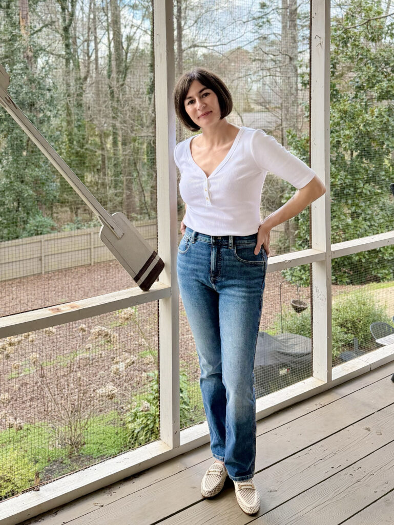 Full-length view of Kelly in her white henley and pencil jeans, hand on hip, leaning against the porch railing in the spring sunshine