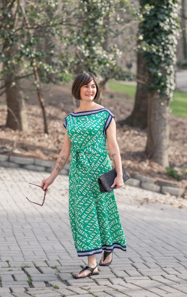 Kelly in Anthropologie Maeve green French Riviera print midi dress, smiling full-length portrait with ivy and spring trees in background, holding black woven clutch — petite maxi dress styling