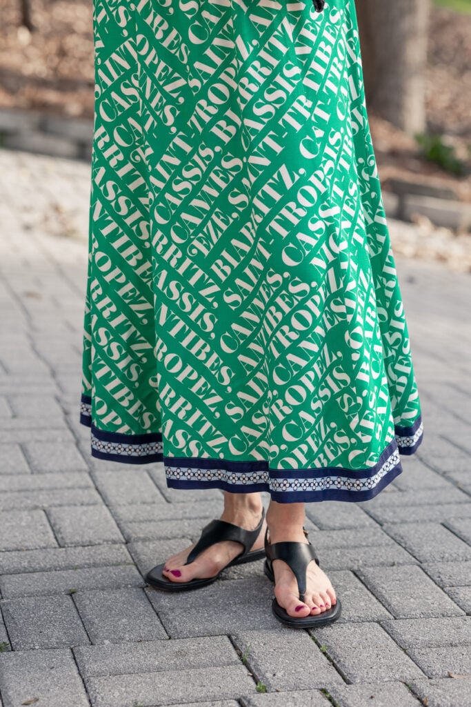 Close-up detail of Anthropologie Maeve Hanky Hem Midi Dress navy border hem and white pattern trim with black covered thong sandals on stone pavers — petite dress hem detail