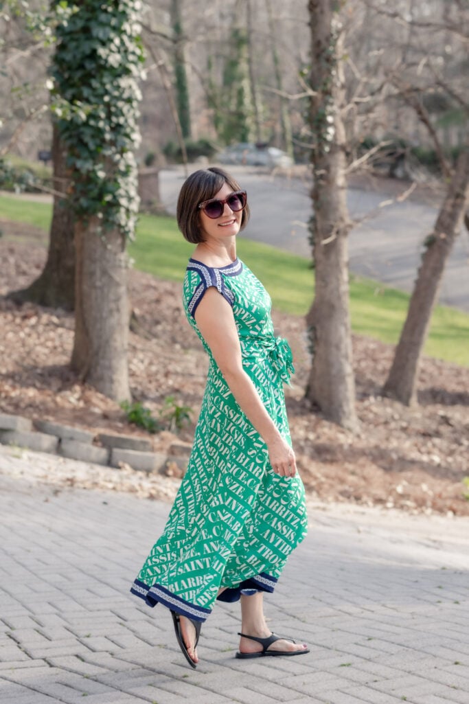 Side view of Kelly in green Anthropologie Maeve Hanky Hem Midi Dress, smiling over shoulder while walking, showing full petite silhouette and navy border hem with black thong sandals