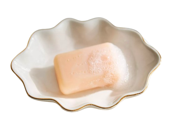 White scalloped-edge ceramic soap dish with delicate gold trim, holding a peach-colored bar of soap with light lather, resting on a folded cream towel.