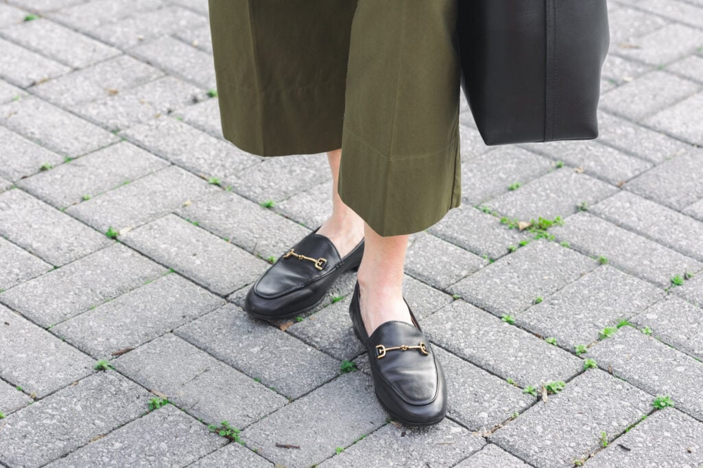Overhead close-up of black Sam Edelman Loraine bit loafers with gold horsebit detail worn with green wide-leg cropped twill pants and black bucket tote — demonstrating the sleek flat shoe silhouette that anchors wide-leg pants on a petite frame