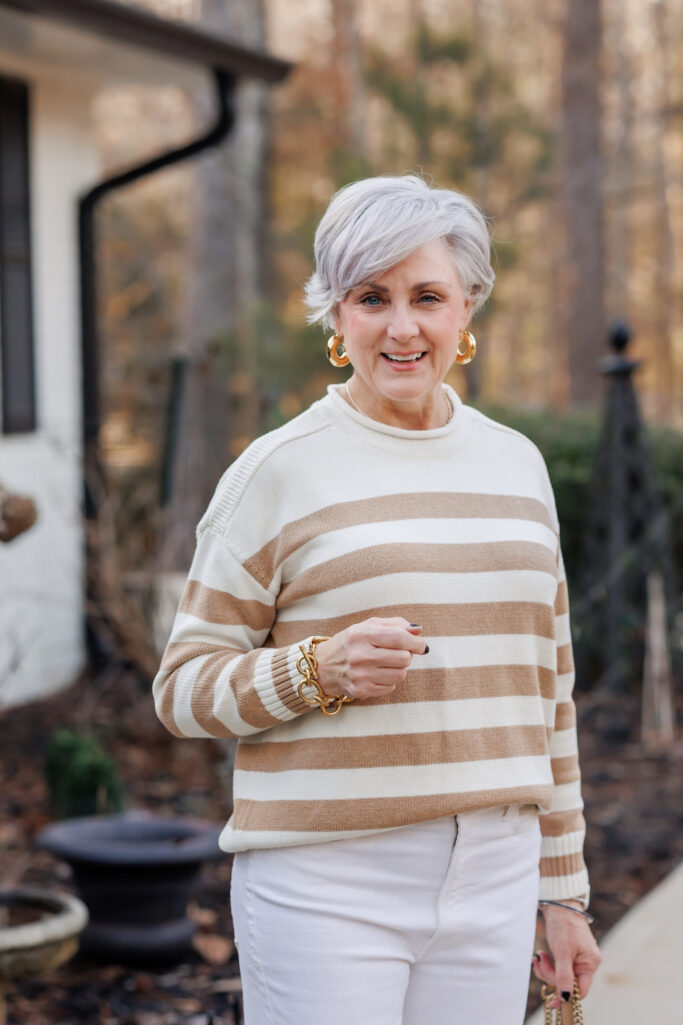 Woman over 50 with silver hair wearing camel and cream striped crewneck sweater with white jeans, chunky gold hoop earrings, gold chain link bracelet