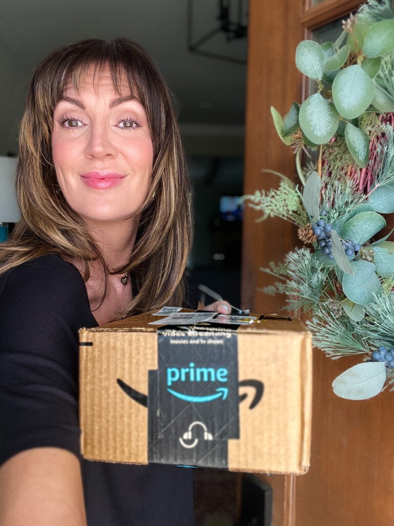 Woman with highlighted brown hair and bangs smiling and holding Amazon Prime shipping box at front door, wearing black ribbed top and small gold heart necklace, greenery wreath with blue berries visible on wooden door behind her