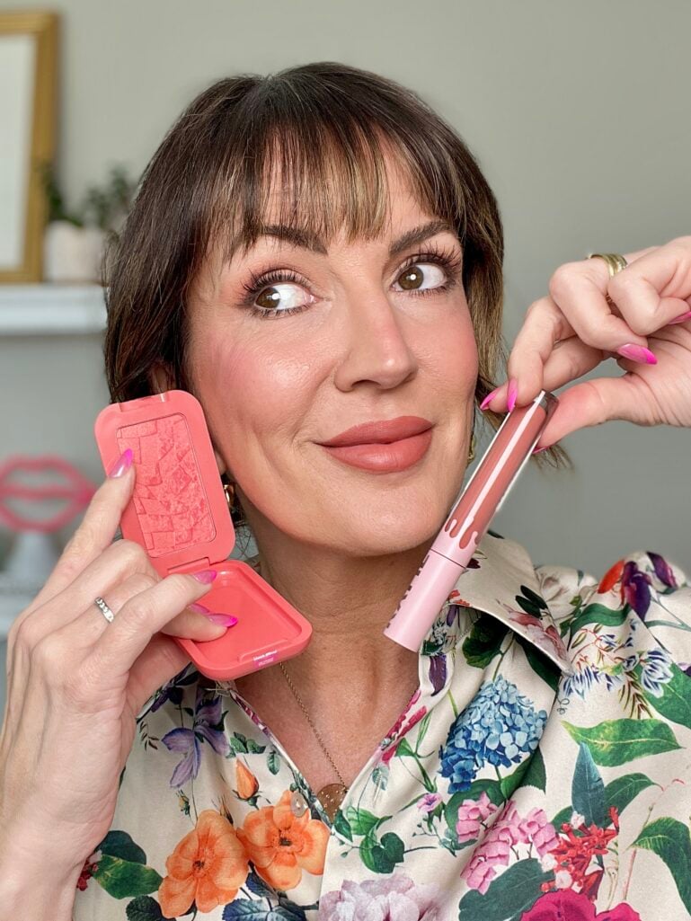 Woman holding open coral NYX Buttermelt blush compact and pink Kylie Cosmetics matte lip product near face, wearing peach-toned makeup look with warm flushed cheeks, floral print blouse, hot pink French tip nails, gold hoop earrings, beauty shelf in background