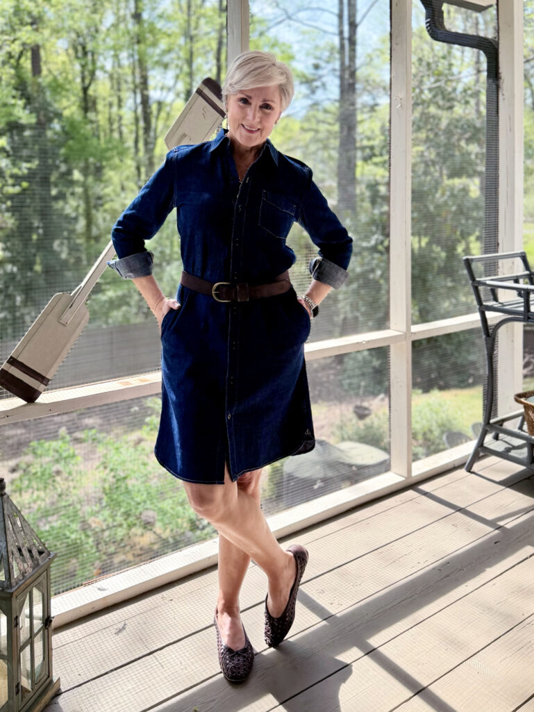 Beth stands on the screened porch in a deep indigo denim shirtdress belted with a dark brown suede belt, sleeves rolled to reveal chambray lining, and woven flats, one hand on her hip and smiling at the camera.
