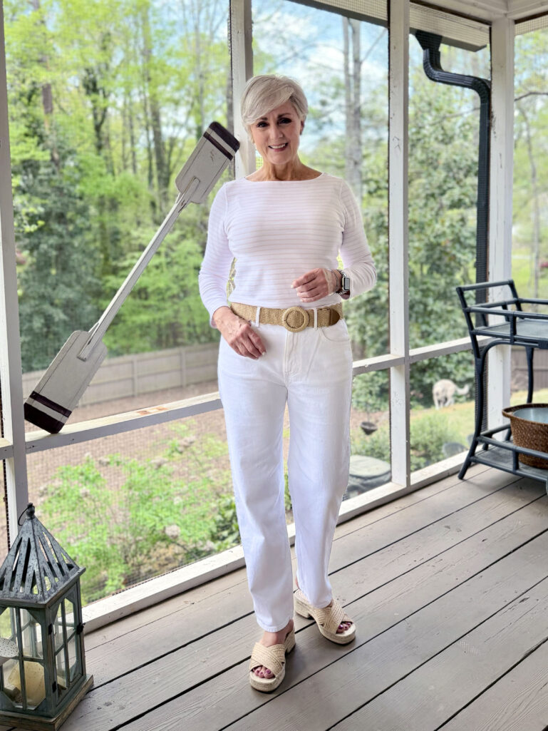 Full-length view of Beth on the screened porch in a white striped long-sleeve tee, white wide-leg jeans, a woven straw belt, and raffia platform slides, smiling warmly with lush green spring trees visible behind her.