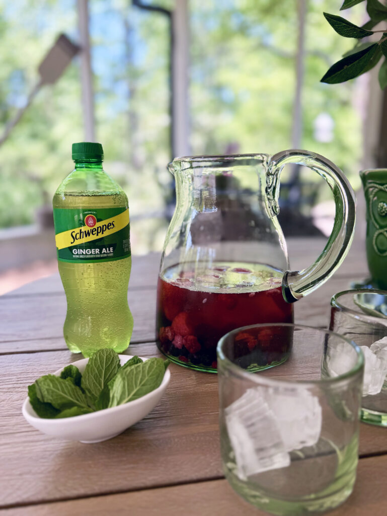 An outdoor wooden table styled with a bottle of Schweppes Ginger Ale, a glass pitcher filled with berry rosé sangria, a small white bowl of fresh mint leaves, and two clear rocks glasses with ice, lush green trees visible through the windows in the background.