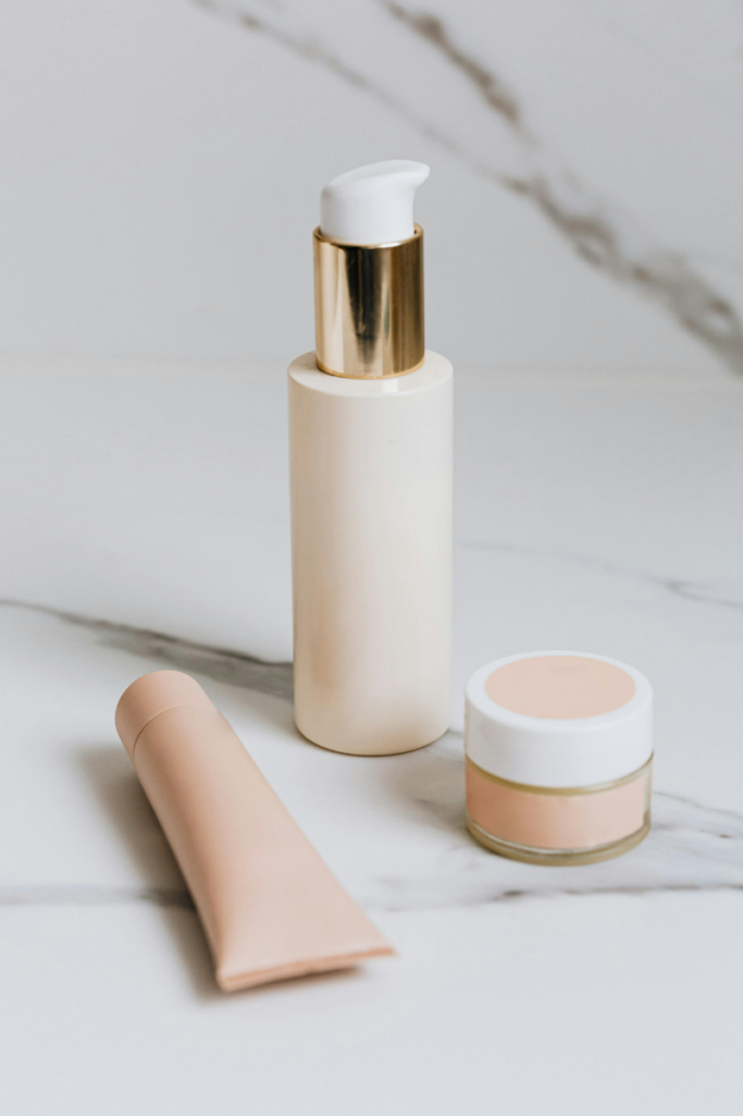 Minimalist skincare products including a cream pump bottle, tube, and small jar in soft neutral tones, styled on a marble surface for a clean, modern beauty routine aesthetic.