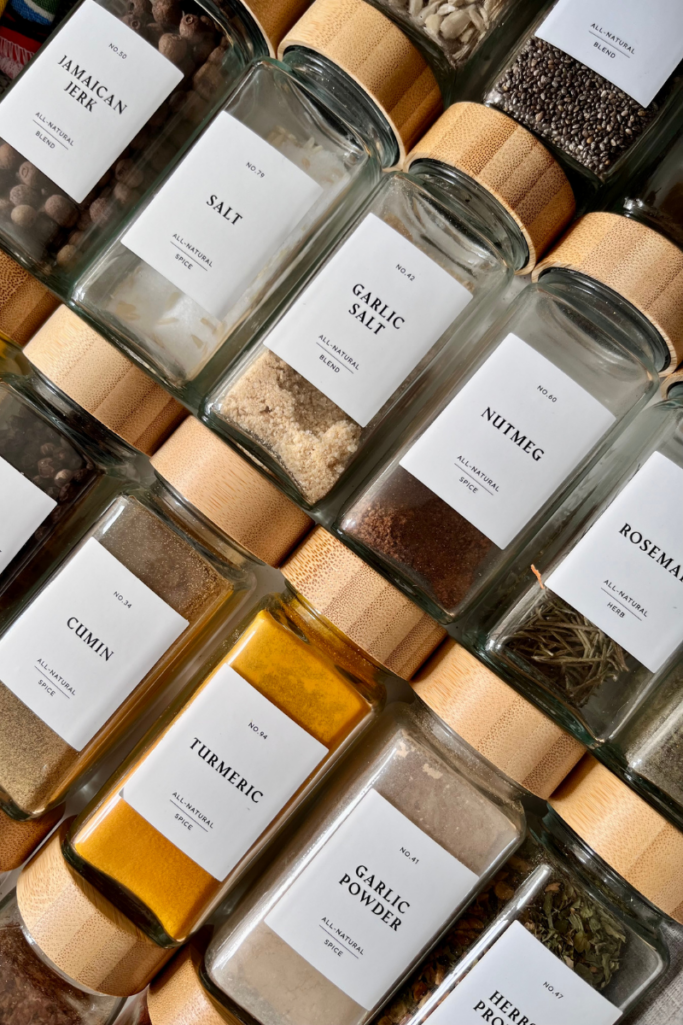 Overhead view of neatly arranged glass spice jars with bamboo lids, labeled with spices like turmeric, cumin, garlic powder, and rosemary for an organized, minimalist kitchen aesthetic.