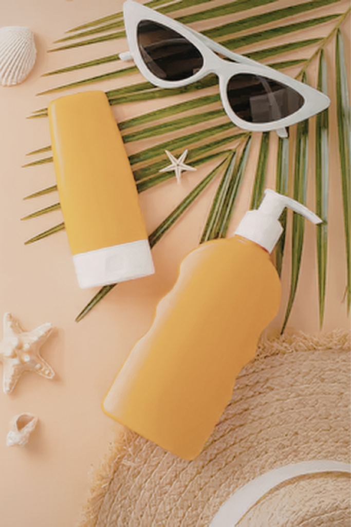 Summer flat lay with yellow sunscreen bottles, white cat-eye sunglasses, palm frond, woven straw hat, and seashell accents on a sandy beige background.