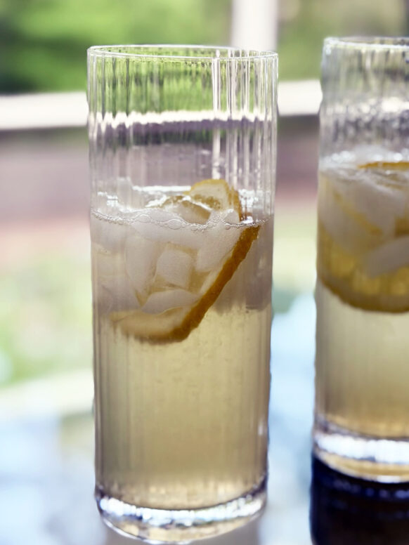 Two Elderflower Collins cocktails in tall ribbed highball glasses sit on a glass table, each garnished with a lemon twist and ice, glowing pale golden in the spring light with soft green bokeh in the background.