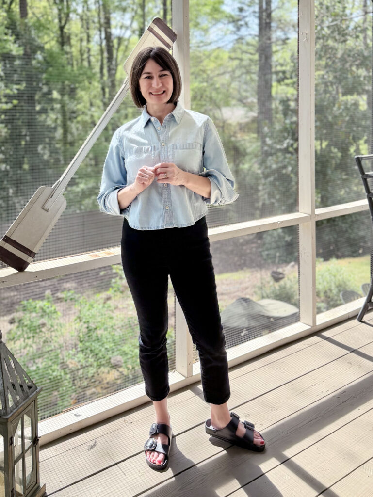 Kelly stands on the screened porch in a light chambray cropped button-front top and black slim ankle pants, hands clasped in front of her, smiling softly at the camera.