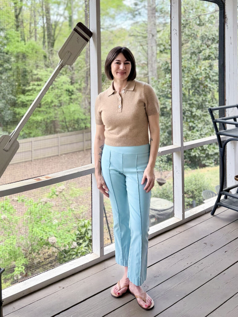 Full-length view of Kelly on the screened porch in a camel polo sweater, sky blue J.McLaughlin Ivy Pants, and easy summer flip-flops, standing relaxed with hands at her sides and smiling at the camera.