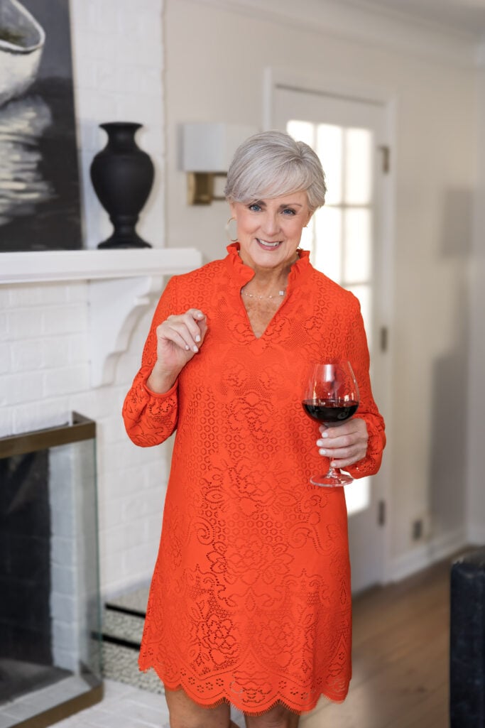 Woman over 50 with silver hair wearing a bold orange lace shift dress with a ruffled mock neckline and scalloped hem, holding a glass of red wine indoors β an example of how to wear orange over 50 with confidence for an evening occasion or entertaining at home.