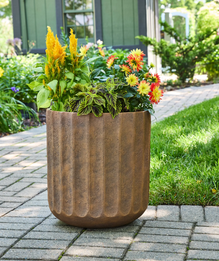 Large textured terracotta-style planter filled with colorful flowers and greenery, styled on a stone patio near a garden walkway.