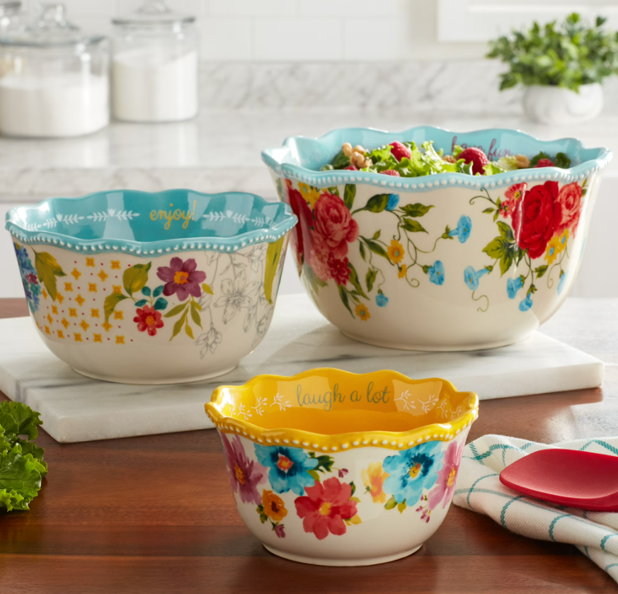 Three-piece Pioneer Woman Sweet Rose ceramic serving bowl set featuring scalloped edges, vibrant floral patterns, and colorful interiors, displayed on a kitchen counter with salad and serving utensils.