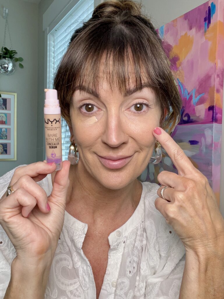 Close-up of hand holding two L'Oreal Paris Le Shadow Stick eyeshadow crayons — one in champagne shimmer and one in warm taupe brown shade labeled Celestial Taupe — pink manicure visible, mirror reflection and makeup products softly blurred in background